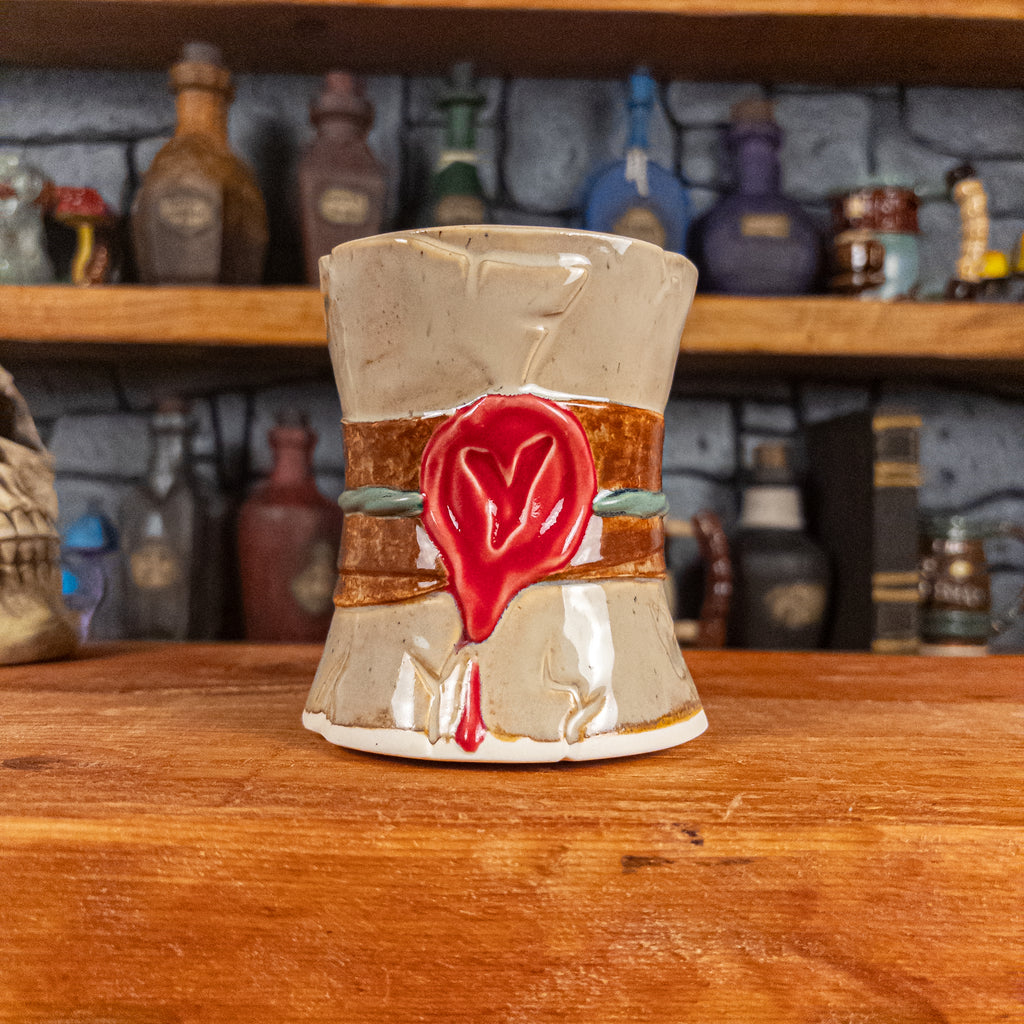 Front view of a handmade Scroll Mug designed to look like an old rolled parchment with a sandy base, a brown ribbon, a greenish-blue cord and a red wax seal featuring a vampire emblem.