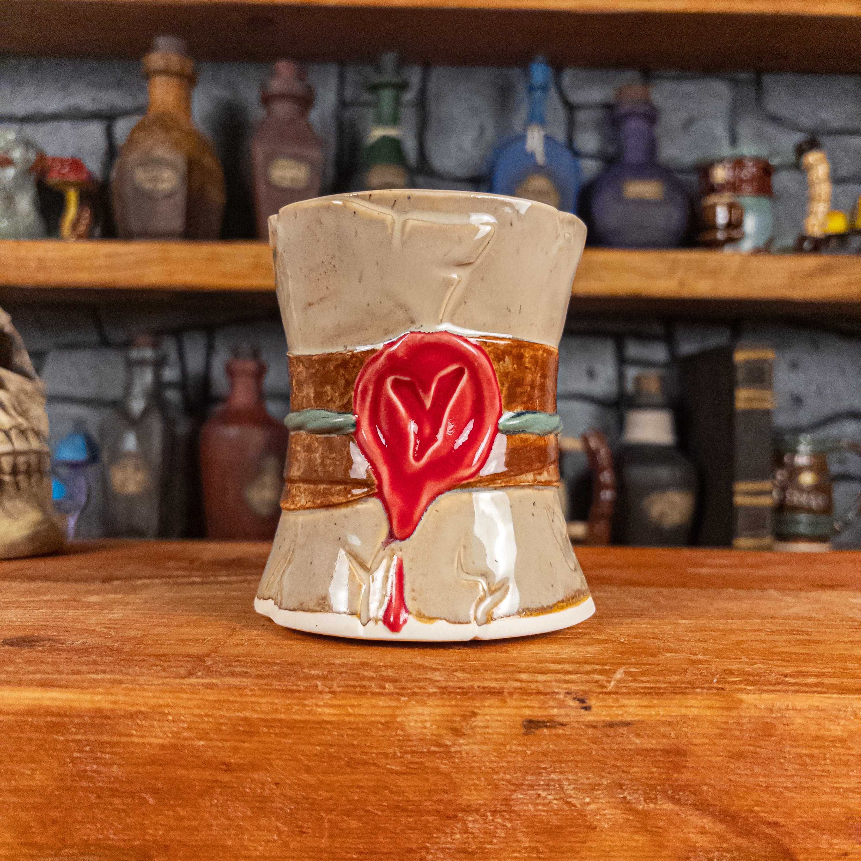 Front view of a handmade Scroll Mug designed to look like an old rolled parchment with a sandy base, a brown ribbon, a greenish-blue cord and a red wax seal featuring a vampire emblem.