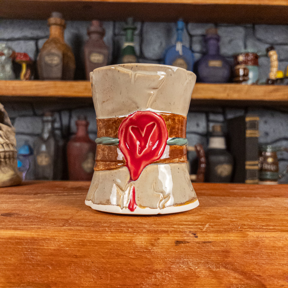 Front view of a handmade Scroll Mug designed to look like an old rolled parchment with a sandy base, a brown ribbon, a greenish-blue cord and a red wax seal featuring a vampire emblem.