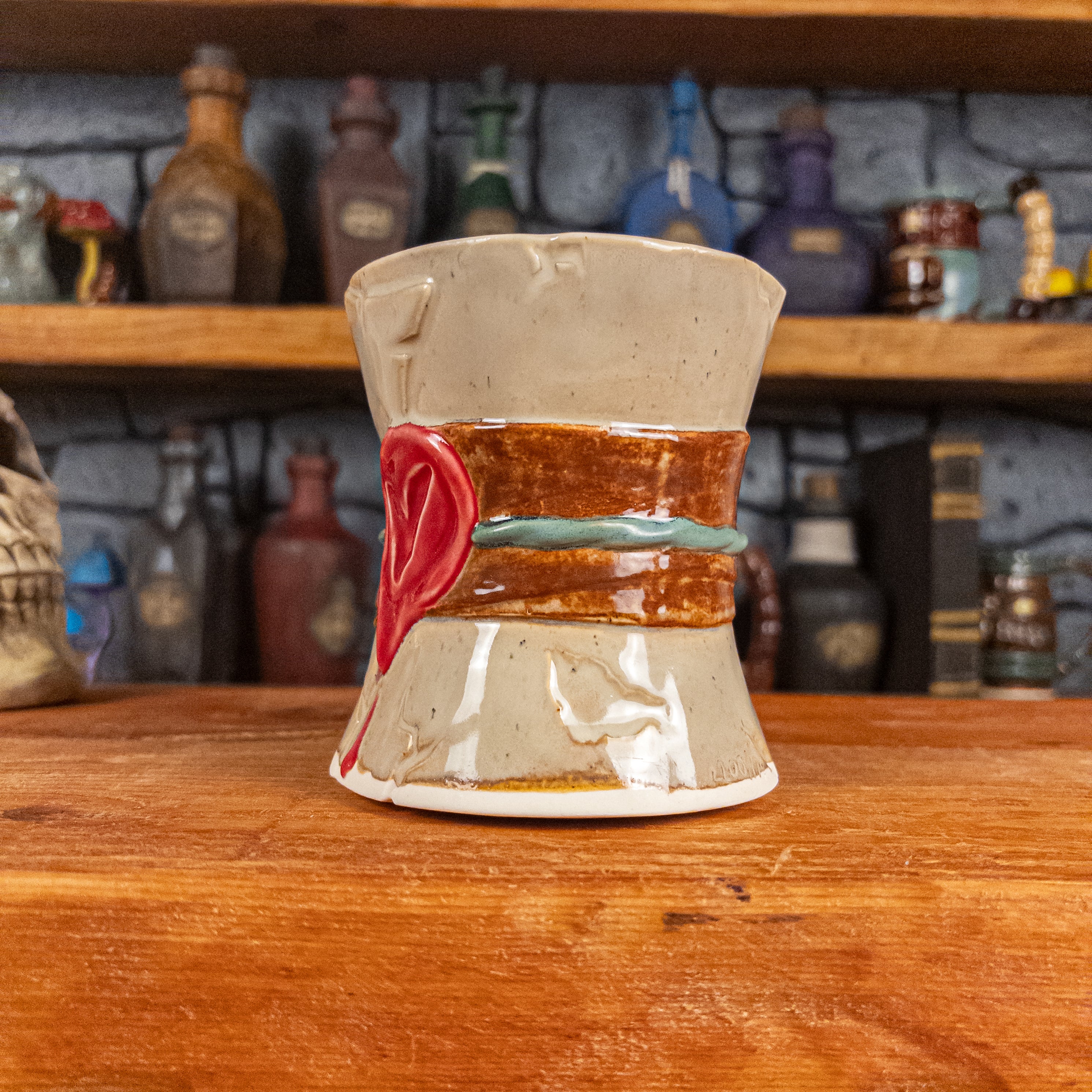 Three-quarter right side view of a handmade Scroll Mug designed to look like an old rolled parchment with a sandy base, a brown ribbon, a greenish-blue cord and a red wax seal featuring a vampire emblem.