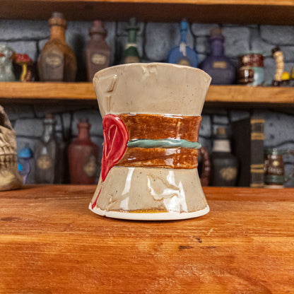 Three-quarter right side view of a handmade Scroll Mug designed to look like an old rolled parchment with a sandy base, a brown ribbon, a greenish-blue cord and a red wax seal featuring a vampire emblem.