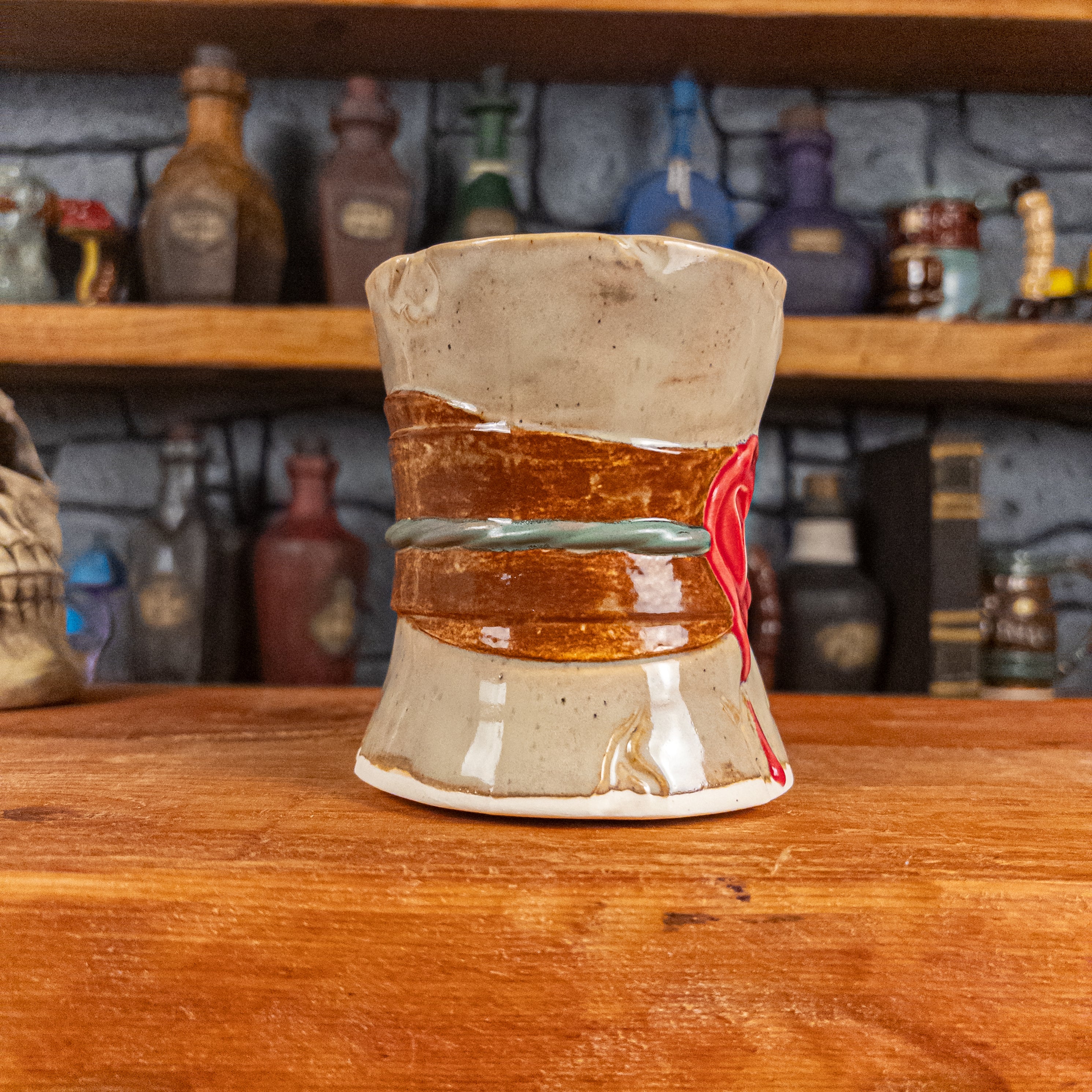 Three-quarter left side view of a handmade Scroll Mug designed to look like an old rolled parchment with a sandy base, a brown ribbon, a greenish-blue cord and a red wax seal featuring a vampire emblem.
