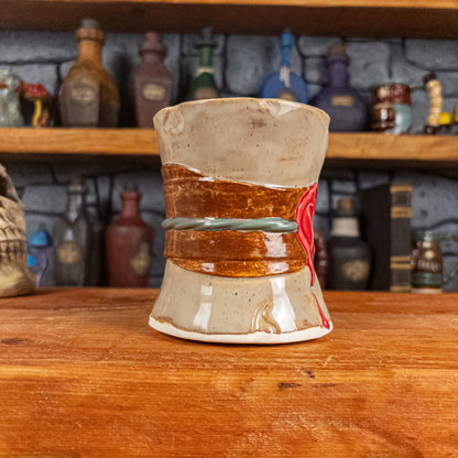 Three-quarter left side view of a handmade Scroll Mug designed to look like an old rolled parchment with a sandy base, a brown ribbon, a greenish-blue cord and a red wax seal featuring a vampire emblem.