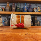 Side view of a handmade Scroll Mug laying down, designed to look like an old rolled parchment with a sandy base, a brown ribbon, a greenish-blue cord and a red wax seal featuring a vampire emblem.