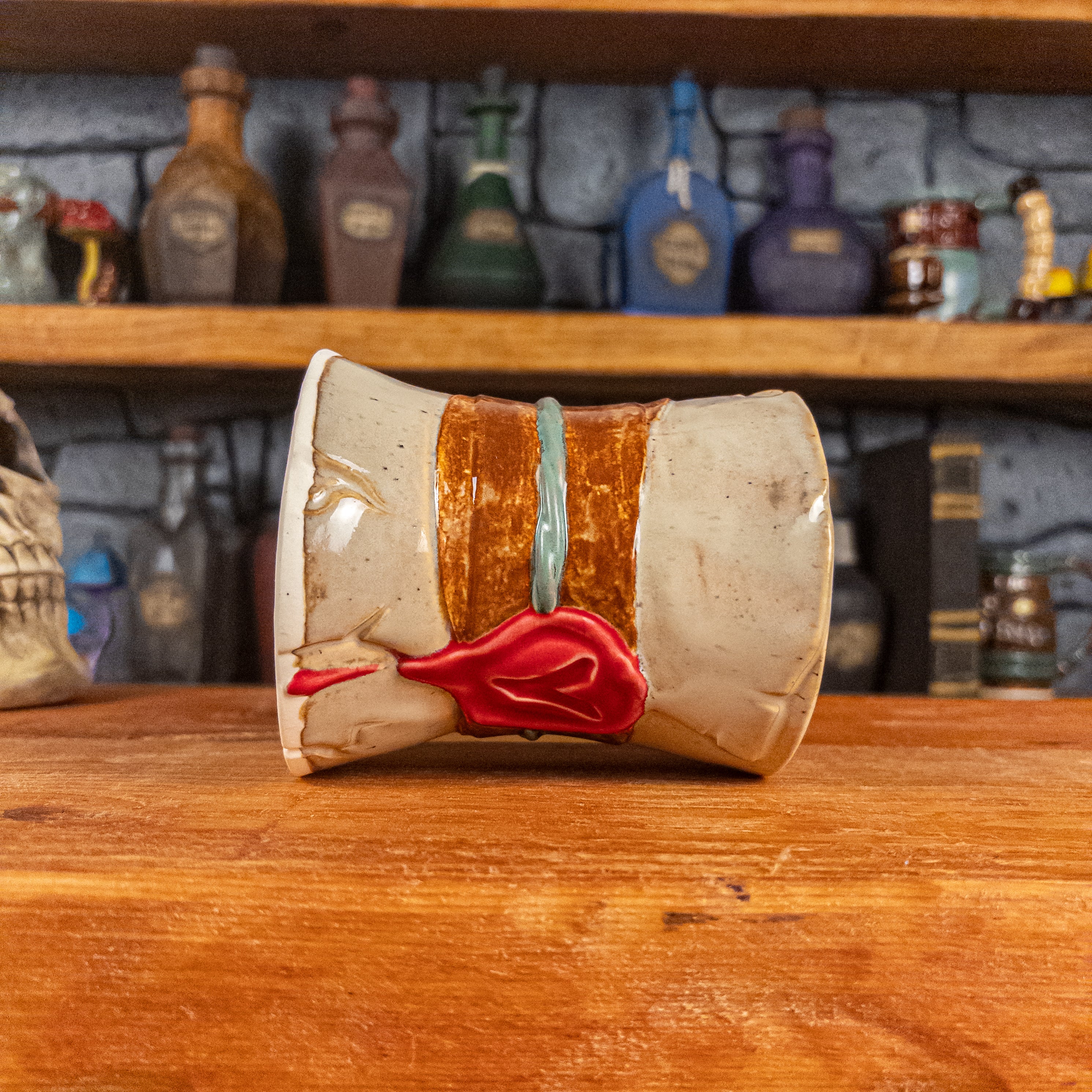 Side view of a handmade Scroll Mug laying down, designed to look like an old rolled parchment with a sandy base, a brown ribbon, a greenish-blue cord and a red wax seal featuring a vampire emblem.
