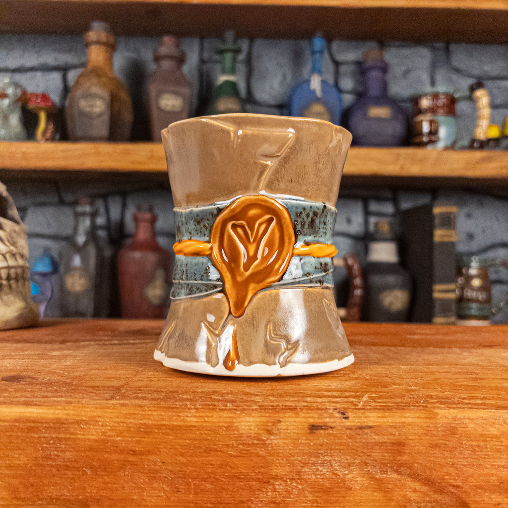 Front view of a handmade Scroll Mug designed to look like an old rolled parchment with a brown base, a speckled blue ribbon, an orange cord and a brown wax seal featuring a vampire emblem.