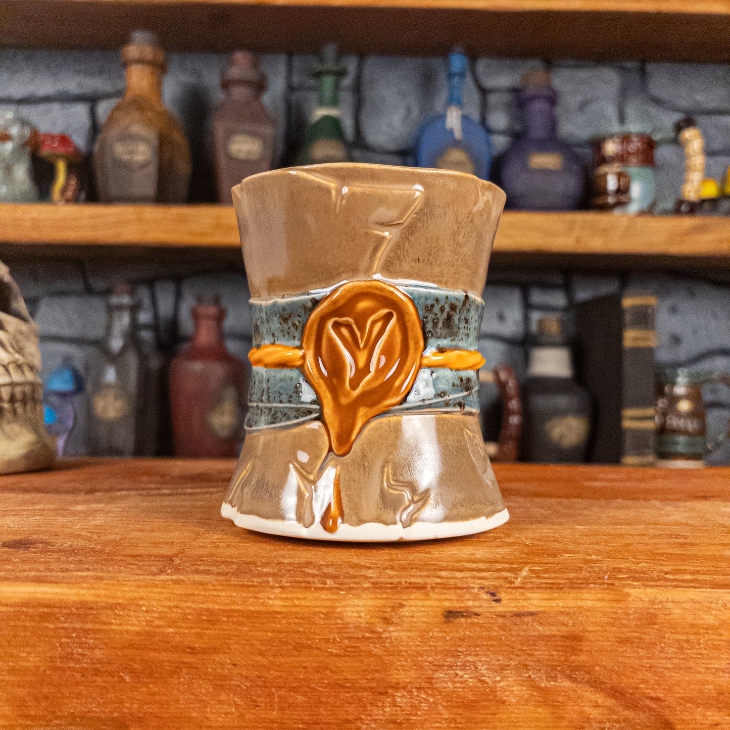 Front view of a handmade Scroll Mug designed to look like an old rolled parchment with a brown base, a speckled blue ribbon, an orange cord and a brown wax seal featuring a vampire emblem.