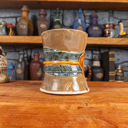 Three-quarter left side view of a handmade Scroll Mug designed to look like an old rolled parchment with a brown base, a speckled blue ribbon, an orange cord and a brown wax seal featuring a vampire emblem.