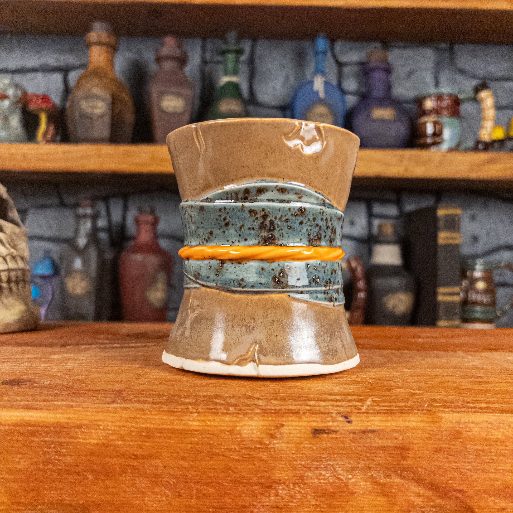 Back view of a handmade Scroll Mug designed to look like an old rolled parchment with a brown base, a speckled blue ribbon and an orange cord.