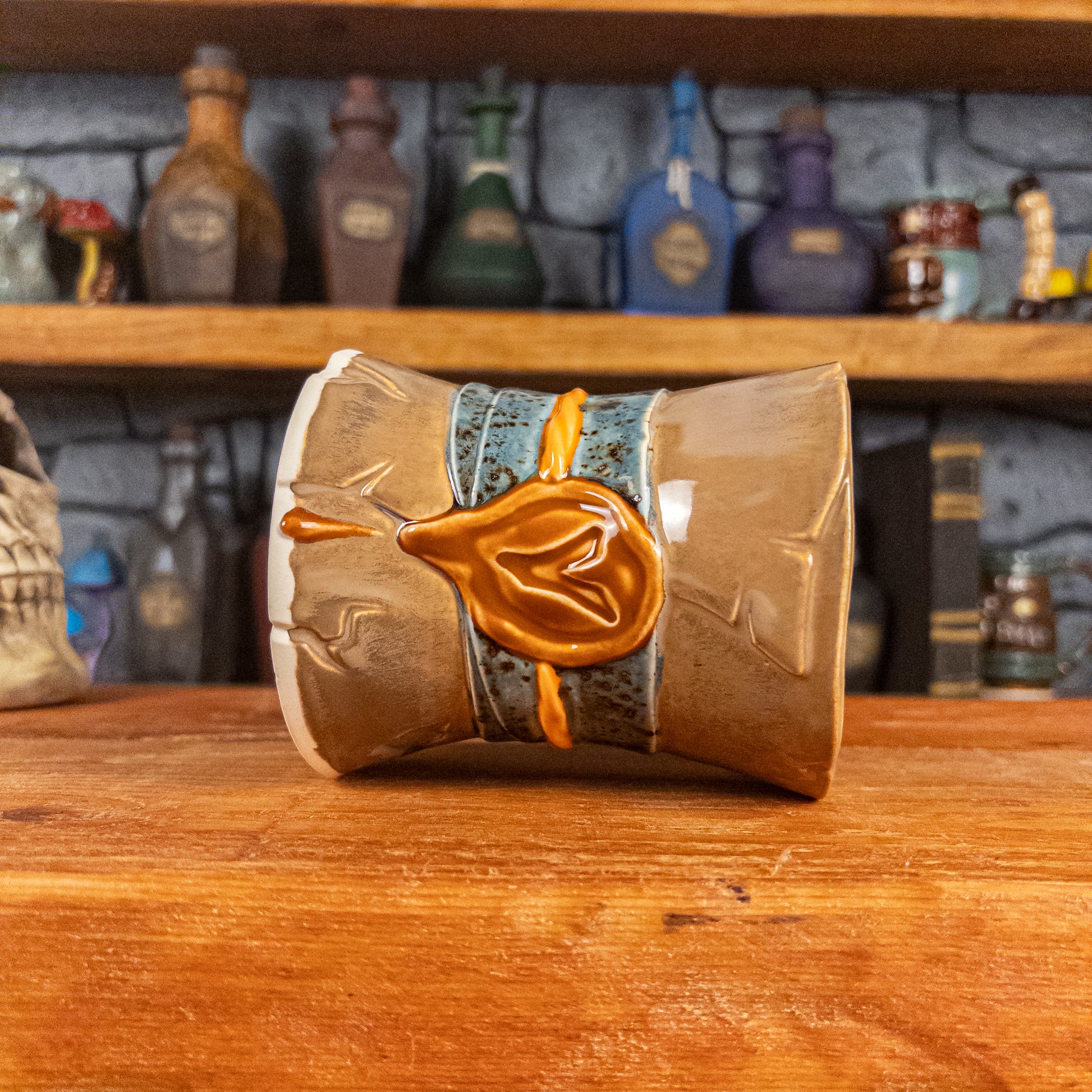 Side view of a handmade Scroll Mug laying down, designed to look like an old rolled parchment with a brown base, a speckled blue ribbon, an orange cord and a brown wax seal featuring a vampire emblem.