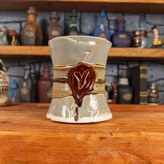 Front view of a handmade Scroll Mug designed to look like an old rolled parchment with a light grey base, a light brown ribbon, a dark brown cord and a dark red wax seal featuring a vampire emblem.