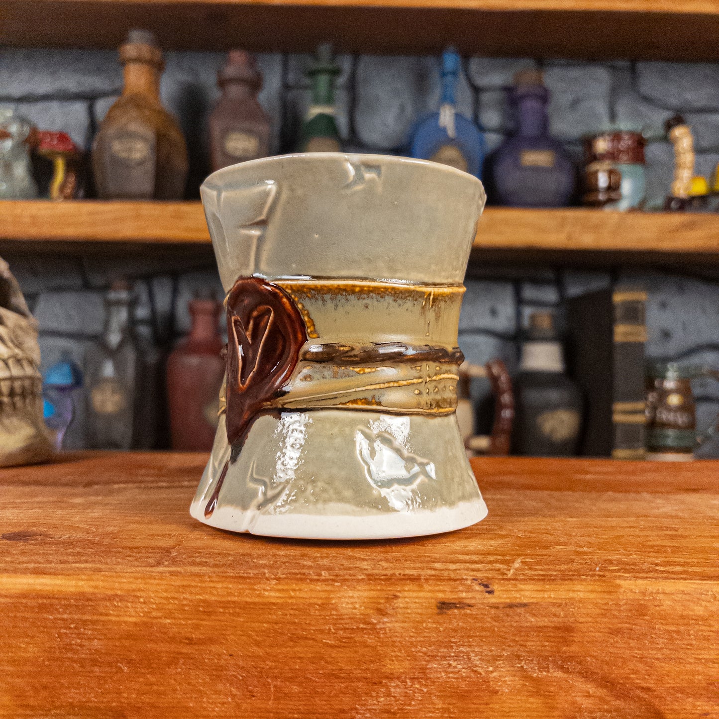Three-quarter right side view of a handmade Scroll Mug designed to look like an old rolled parchment with a light grey base, a light brown ribbon, a dark brown cord and a dark red wax seal featuring a vampire emblem.