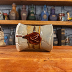 Side view of a handmade Scroll Mug laying down, designed to look like an old rolled parchment with a light grey base, a light brown ribbon, a dark brown cord and a dark red wax seal featuring a vampire emblem.