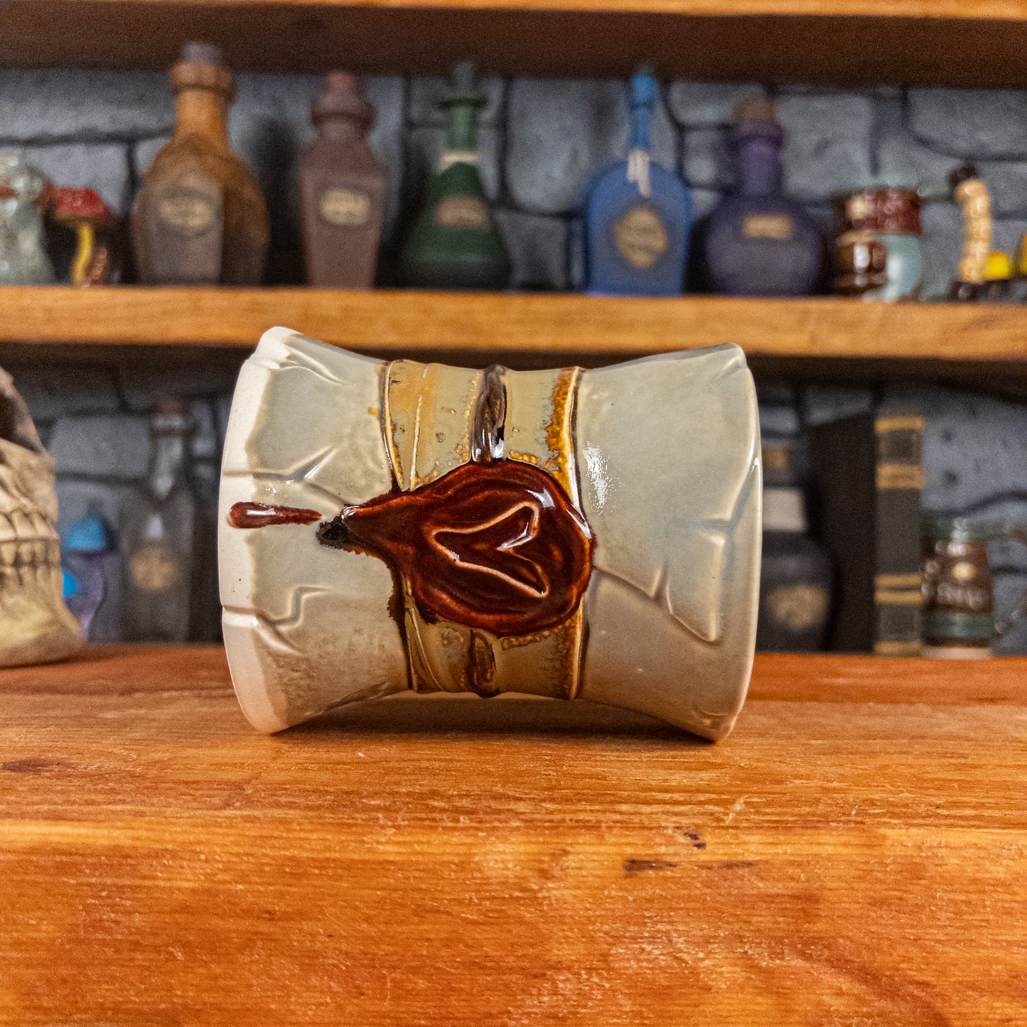 Side view of a handmade Scroll Mug laying down, designed to look like an old rolled parchment with a light grey base, a light brown ribbon, a dark brown cord and a dark red wax seal featuring a vampire emblem.