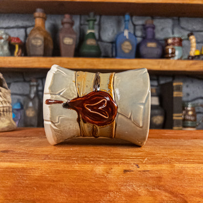 Side view of a handmade Scroll Mug laying down, designed to look like an old rolled parchment with a light grey base, a light brown ribbon, a dark brown cord and a dark red wax seal featuring a vampire emblem.