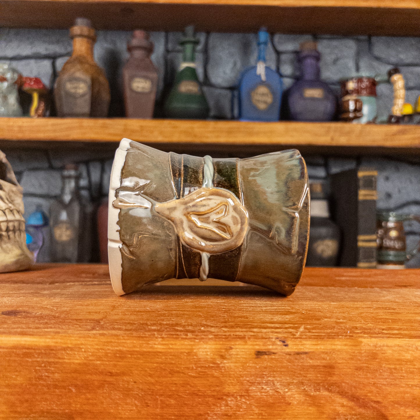 Side view of a handmade Scroll Mug laying down, designed to look like an old rolled parchment with a brown base, a brown-green ribbon, a light green cord and a beige wax seal featuring a vampire emblem.