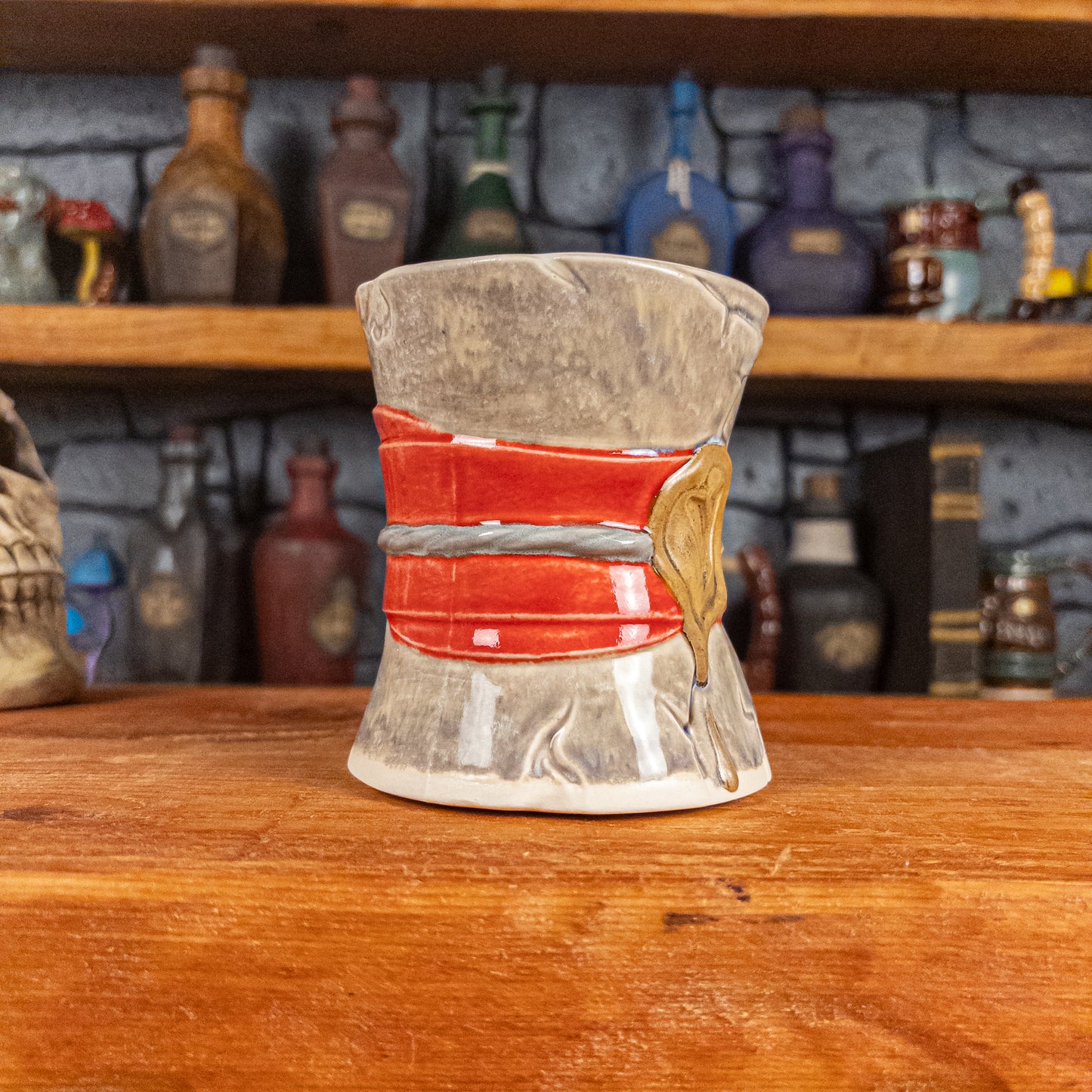 Three-quarter left side view of a handmade Scroll Mug designed to look like an old rolled parchment with a grey base, a red ribbon, a silver cord and a golden wax seal featuring a vampire emblem.