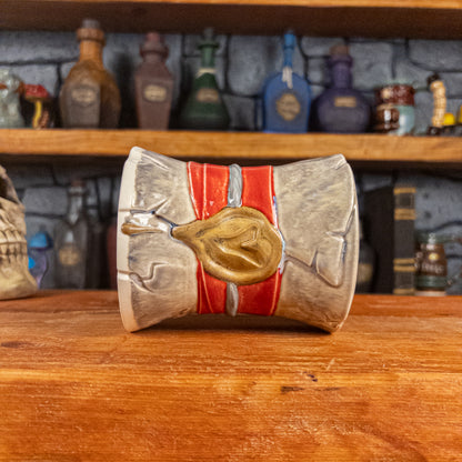 Side view of a handmade Scroll Mug laying down, designed to look like an old rolled parchment with a grey base, a red ribbon, a silver cord and a golden wax seal featuring a vampire emblem.