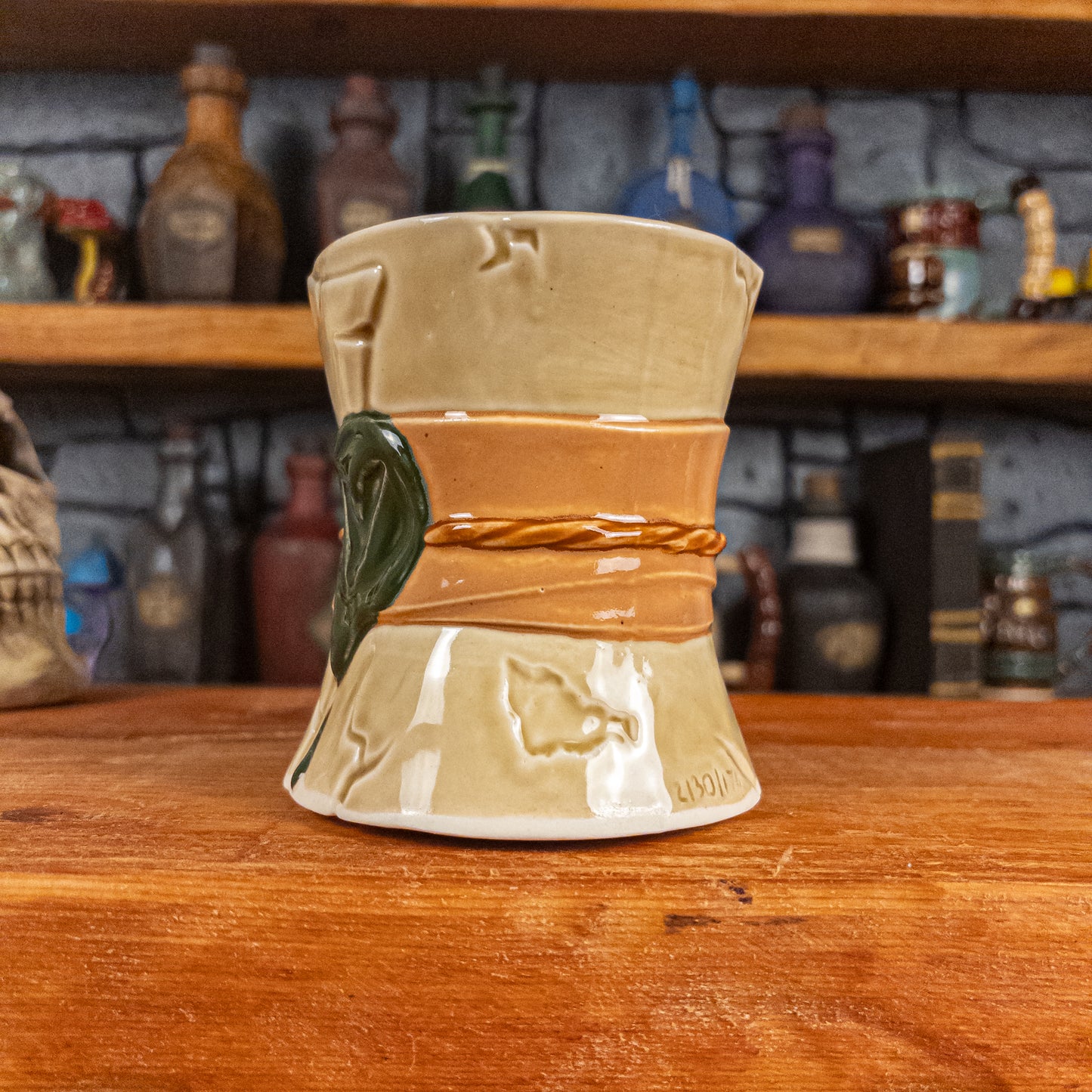 Three-quarter right side view of a handmade Scroll Mug designed to look like an old rolled parchment with a light yellow base, a light orange ribbon, a light brown cord and a dark green wax seal featuring a vampire emblem.