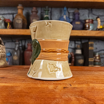 Three-quarter right side view of a handmade Scroll Mug designed to look like an old rolled parchment with a light yellow base, a light orange ribbon, a light brown cord and a dark green wax seal featuring a vampire emblem.