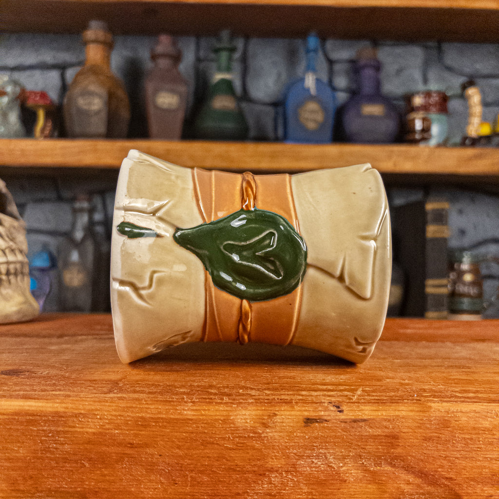 Side view of a handmade Scroll Mug laying down, designed to look like an old rolled parchment with a light-yellow base, a light orange ribbon, a light brown cord and a dark green wax seal featuring a vampire emblem.