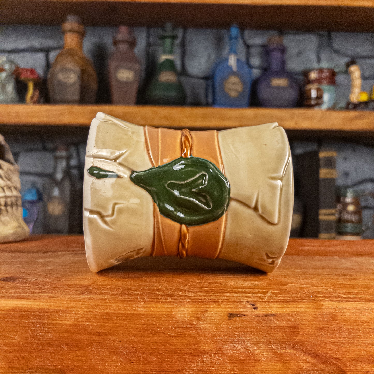 Side view of a handmade Scroll Mug laying down, designed to look like an old rolled parchment with a light-yellow base, a light orange ribbon, a light brown cord and a dark green wax seal featuring a vampire emblem.