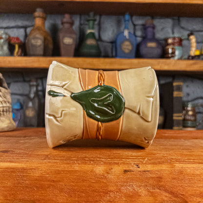Side view of a handmade Scroll Mug laying down, designed to look like an old rolled parchment with a light-yellow base, a light orange ribbon, a light brown cord and a dark green wax seal featuring a vampire emblem.