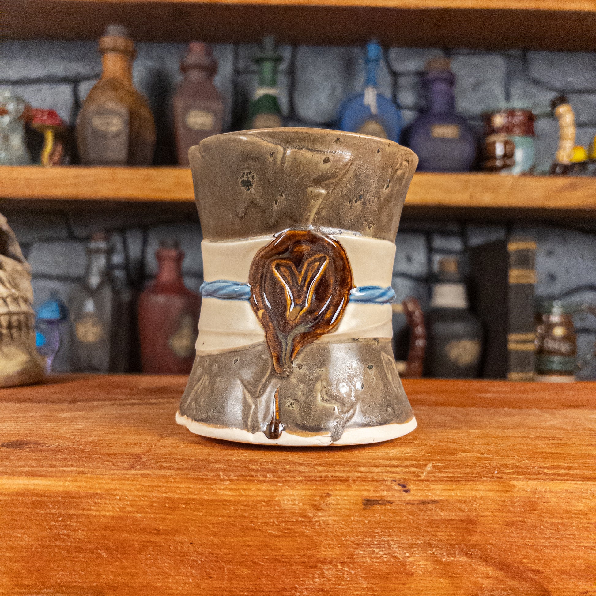Front view of a handmade Scroll Mug designed to look like an old rolled parchment with a speckled dark brown base, an alabaster ribbon, a light blue cord and a reddish-brown wax seal featuring a vampire emblem.