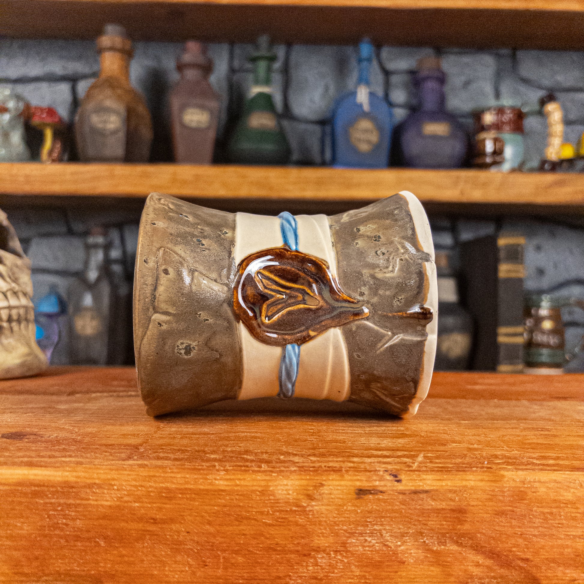 Side view of a handmade Scroll Mug laying down, designed to look like an old rolled parchment with a speckled dark brown base, an alabaster ribbon, a light blue cord and a reddish-brown wax seal featuring a vampire emblem.