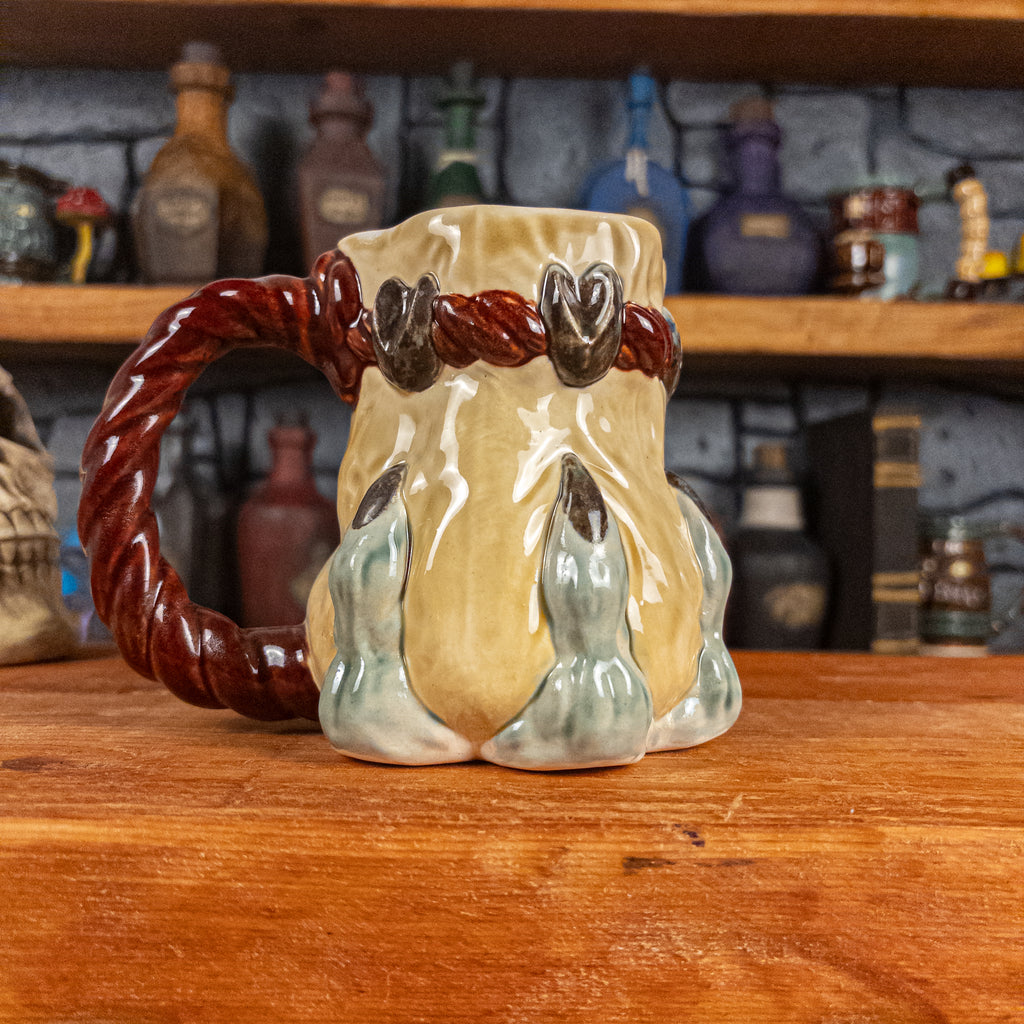 Left side view of a handmade Coin Bag Mug held by a dragon hand with three light green fingers and dark brown nails. The mug has a detailed design featuring a light-yellow coin bag with a dark red braided rope as a handle.