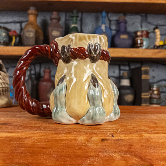 Left side view of a handmade Coin Bag Mug held by a dragon hand with three light green fingers and dark brown nails. The mug has a detailed design featuring a light-yellow coin bag with a dark red braided rope as a handle.