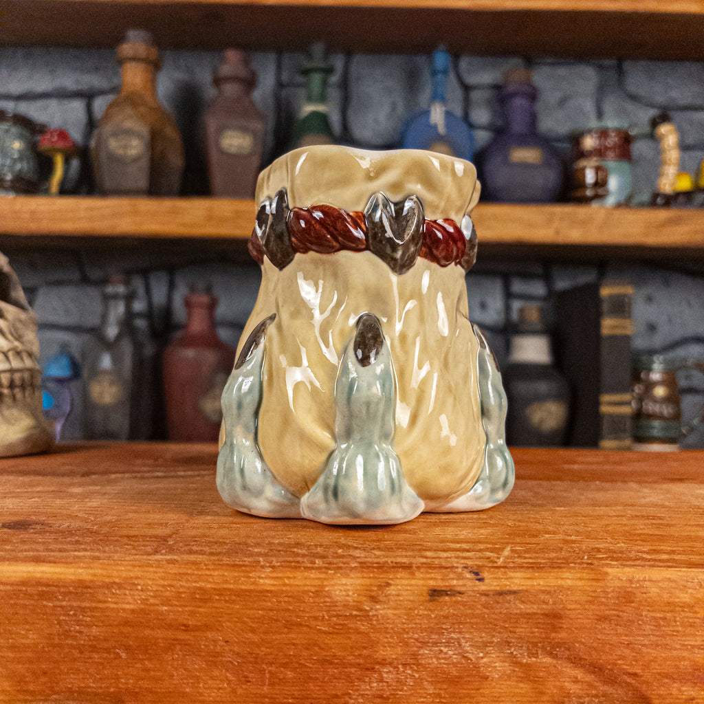 Front view of a handmade Coin Bag Mug held by a dragon hand with three light green fingers and dark brown nails. The mug has a detailed design featuring a light-yellow coin bag.