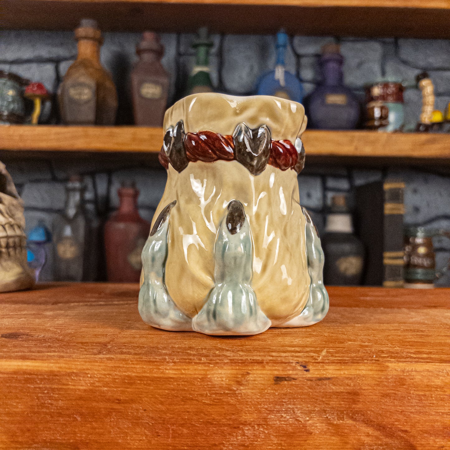 Front view of a handmade Coin Bag Mug held by a dragon hand with three light green fingers and dark brown nails. The mug has a detailed design featuring a light-yellow coin bag.