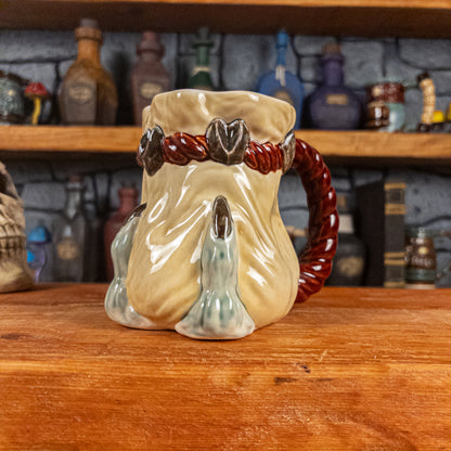 Three-quarter right side view of a handmade Coin Bag Mug held by a dragon hand with two light green fingers and dark brown nails. The mug has a detailed design featuring a light-yellow coin bag with a dark red braided rope as a handle.