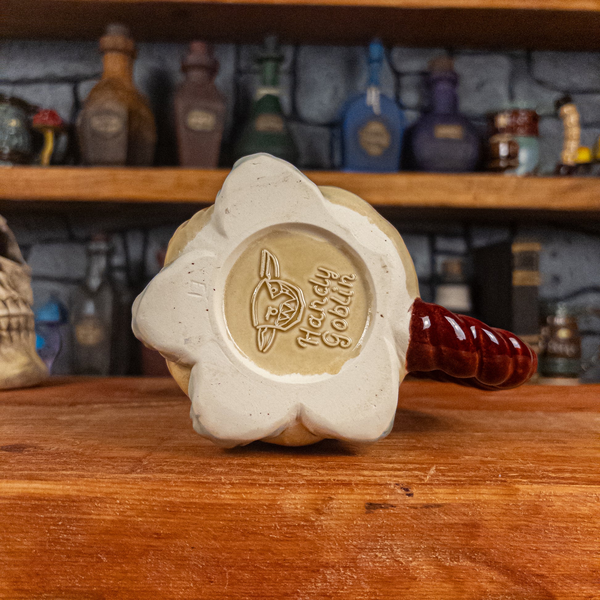 Bottom view of a handmade Coin Bag Mug with the Handy Goblin logo embossed in the centre, featuring a light-yellow base with the visible bottom of the four fingers.