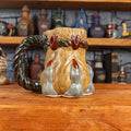 Left side view of a handmade Coin Bag Mug held by a dragon's hand with three light greenish-blue fingers and dark red nails. The mug has a detailed design featuring a golden coin bag with a dark green braided rope as a handle.