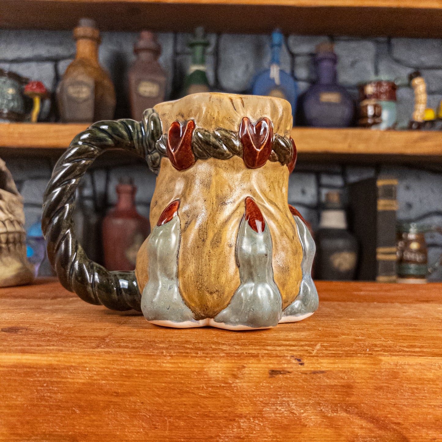 Left side view of a handmade Coin Bag Mug held by a dragon's hand with three light greenish-blue fingers and dark red nails. The mug has a detailed design featuring a golden coin bag with a dark green braided rope as a handle.