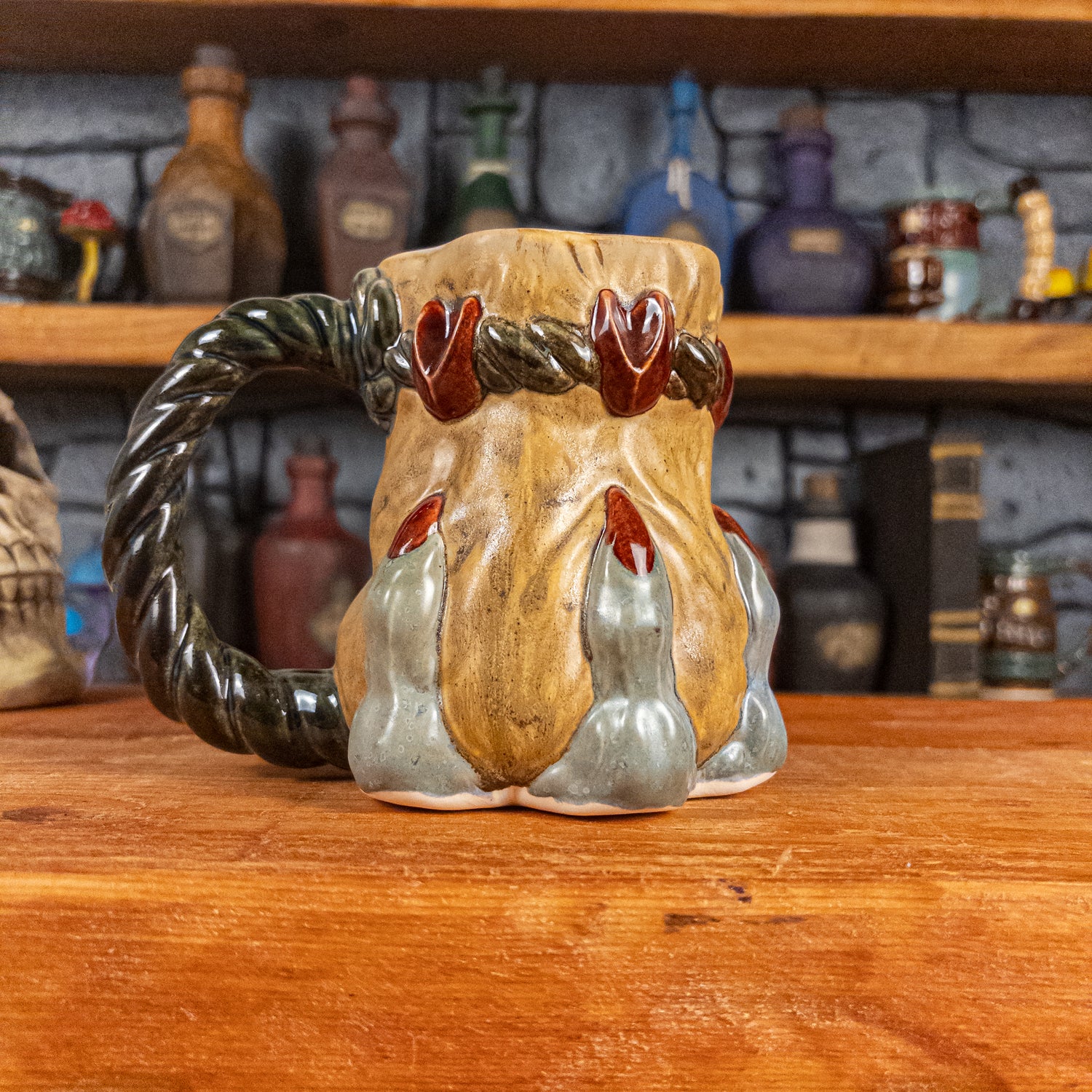 Left side view of a handmade Coin Bag Mug held by a dragon's hand with three light greenish-blue fingers and dark red nails. The mug has a detailed design featuring a golden coin bag with a dark green braided rope as a handle.