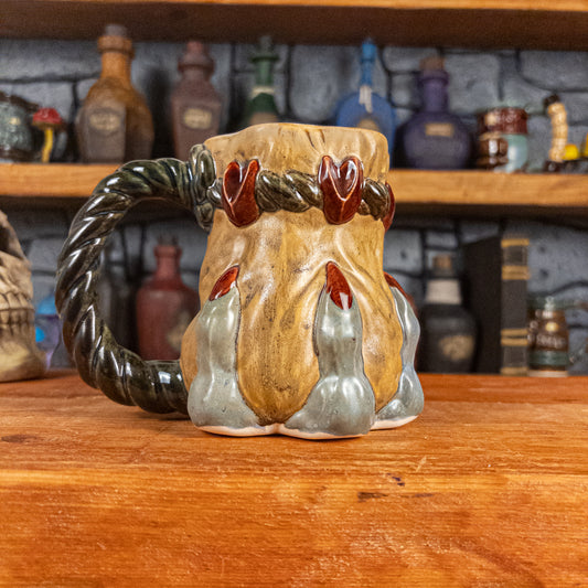 Left side view of a handmade Coin Bag Mug held by a dragon's hand with three light greenish-blue fingers and dark red nails. The mug has a detailed design featuring a golden coin bag with a dark green braided rope as a handle.