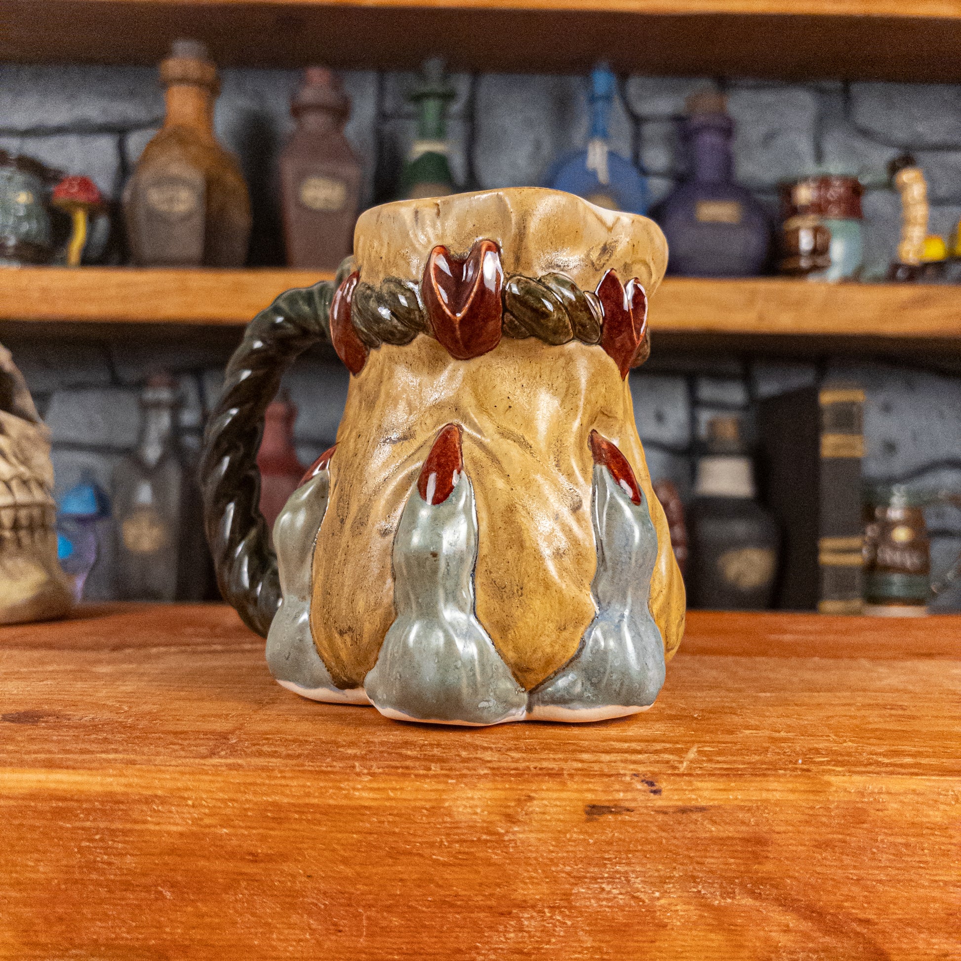 Three-quarter left side view of a handmade Coin Bag Mug held by a dragon's hand with three light greenish-blue fingers and dark red nails. The mug has a detailed design featuring a golden coin bag with a dark green braided rope as a handle.