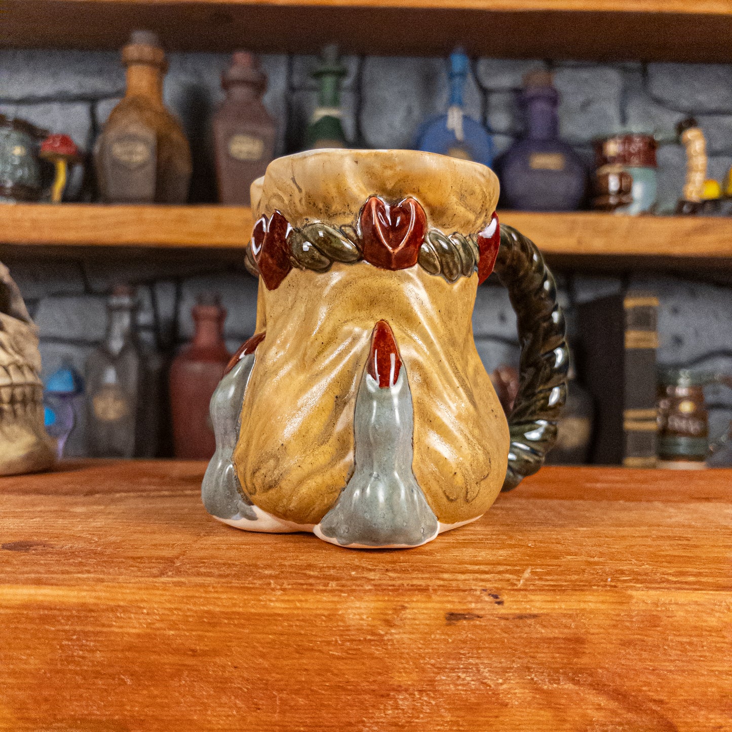 Three-quarter right side view of a handmade Coin Bag Mug held by a dragon's hand with three light greenish-blue fingers and dark red nails. The mug has a detailed design featuring a golden coin bag with a dark green braided rope as a handle.