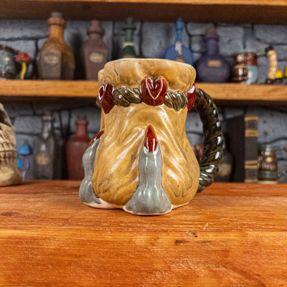 Three-quarter right side view of a handmade Coin Bag Mug held by a dragon's hand with three light greenish-blue fingers and dark red nails. The mug has a detailed design featuring a golden coin bag with a dark green braided rope as a handle.