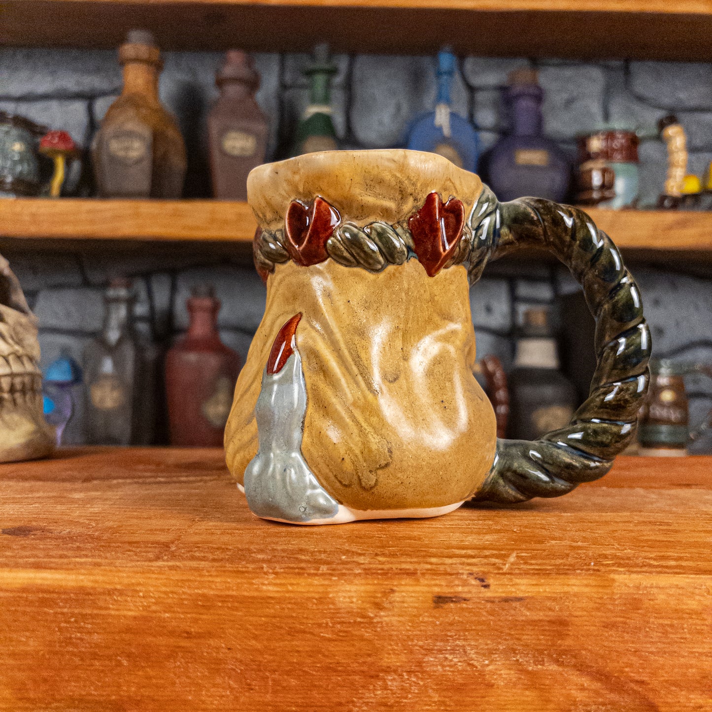 Right side view of a handmade Coin Bag Mug held by a dragon's hand with one light greenish-blue finger and dark red nail. The mug has a detailed design featuring a gold coin bag with a dark green braided rope as a handle.