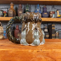 Left side view of a handmade Coin Bag Mug held by a dragon hand with three greenish-blue fingers and dark brown nails. The mug has a detailed design featuring a greenish-brown coin bag with a blue-green braided rope as a handle.