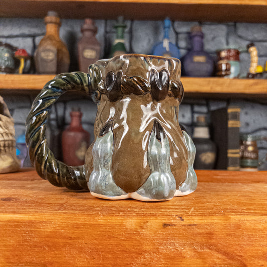 Left side view of a handmade Coin Bag Mug held by a dragon hand with three greenish-blue fingers and dark brown nails. The mug has a detailed design featuring a greenish-brown coin bag with a blue-green braided rope as a handle.