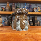 Three-quarter left side view of a handmade Coin Bag Mug held by a dragon hand with three greenish-blue fingers and dark brown nails. The mug has a detailed design featuring a greenish-brown coin bag with a blue-green braided rope handle.