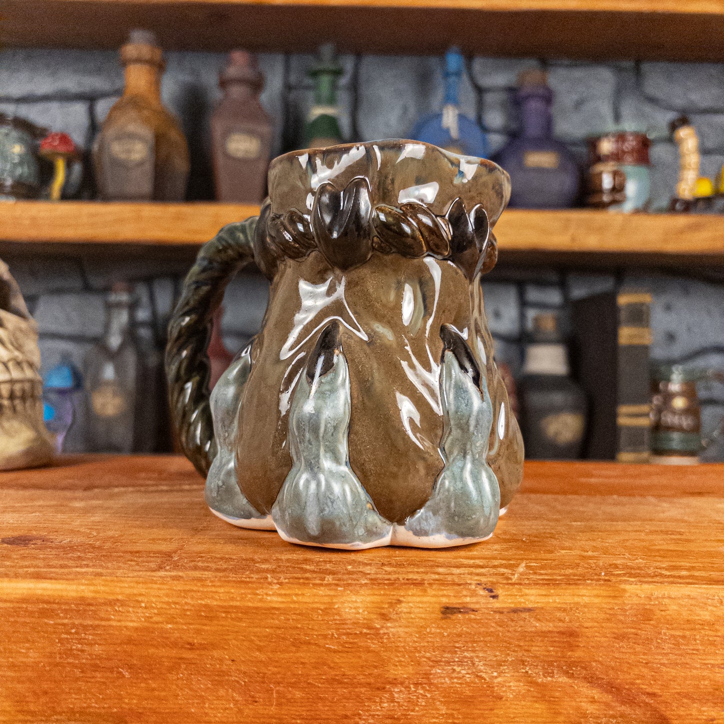 Three-quarter left side view of a handmade Coin Bag Mug held by a dragon hand with three greenish-blue fingers and dark brown nails. The mug has a detailed design featuring a greenish-brown coin bag with a blue-green braided rope handle.