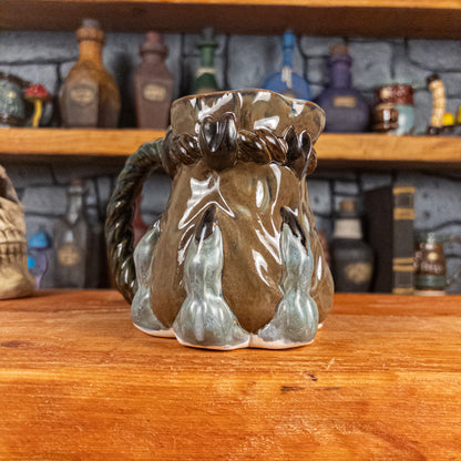 Three-quarter left side view of a handmade Coin Bag Mug held by a dragon hand with three greenish-blue fingers and dark brown nails. The mug has a detailed design featuring a greenish-brown coin bag with a blue-green braided rope handle.
