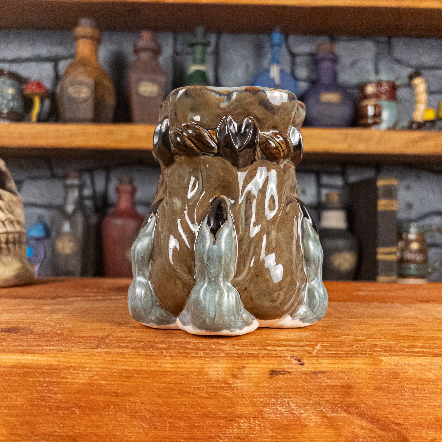 Front view of a handmade Coin Bag Mug held by a dragon hand with three greenish-blue fingers and dark brown nails. The mug has a detailed design featuring a greenish-brown coin bag.
