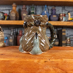 Three-quarter right side view of a handmade Coin Bag Mug held by a dragon hand with two greenish-blue fingers and dark brown nails. The mug has a detailed design featuring a greenish-brown coin bag with a blue-green braided rope handle.